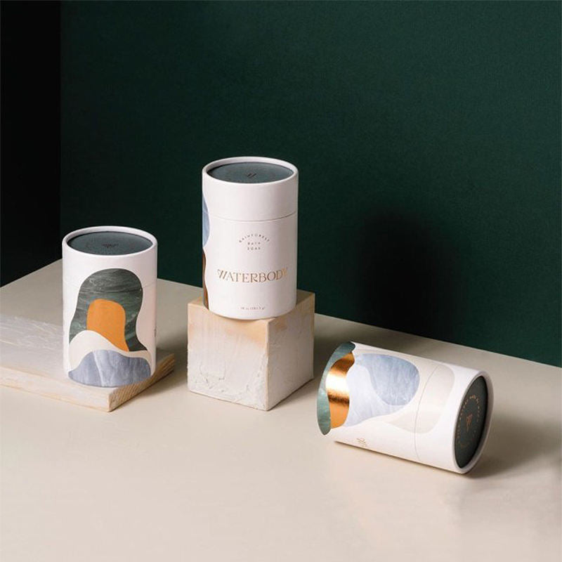 Three artistic cylindrical paper tubes with abstract design elements in earthy tones, displayed against a two-tone dark green and cream background, suggesting a sophisticated, eco-conscious product packaging.