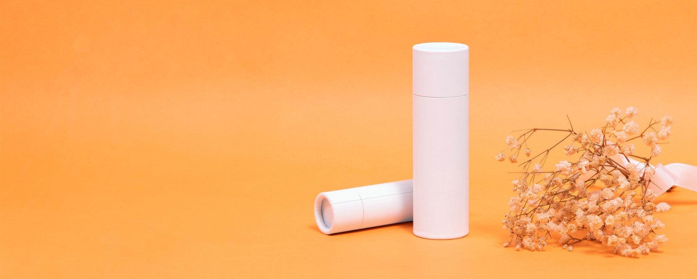 Two white paper tubes standing upright and lying down on an orange backdrop, accompanied by delicate beige dried flowers, presenting a clean and simplistic aesthetic.