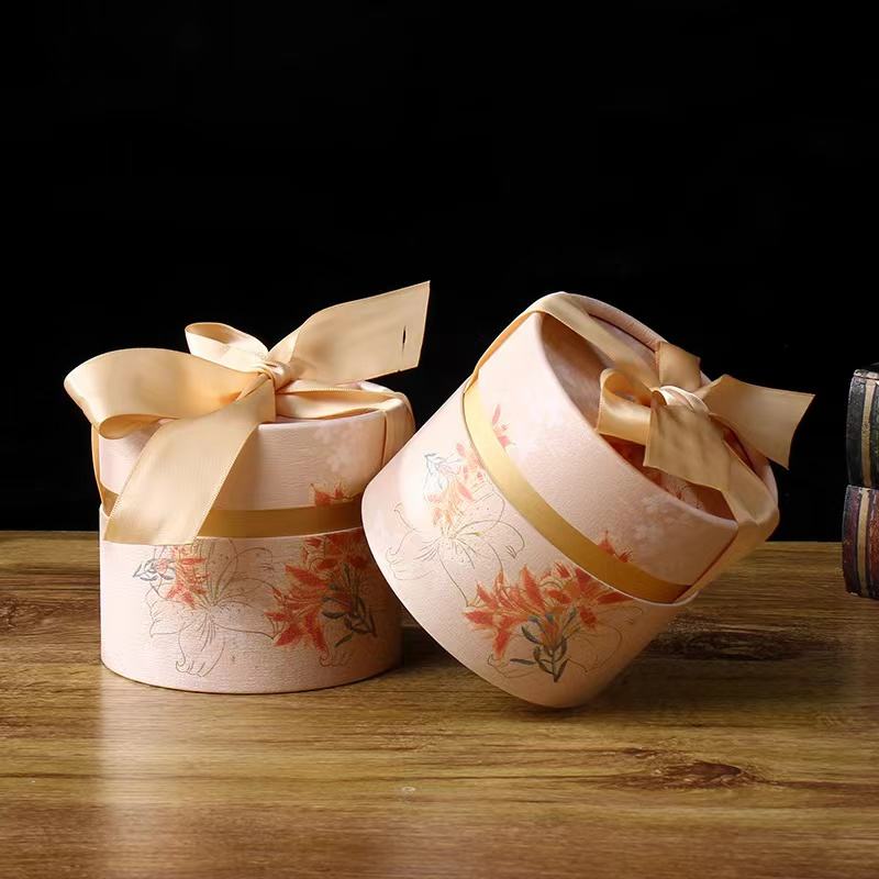 Two elegantly wrapped round paper tubes with floral prints and silky cream ribbons, presented on a wooden surface with a dark backdrop, suggesting a luxurious gift.