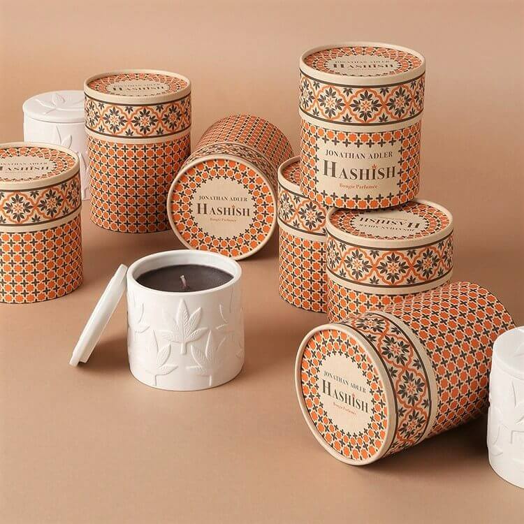 Decorative paper tubes with intricate orange patterns are clustered together, one revealing a candle, against a cohesive tan background.