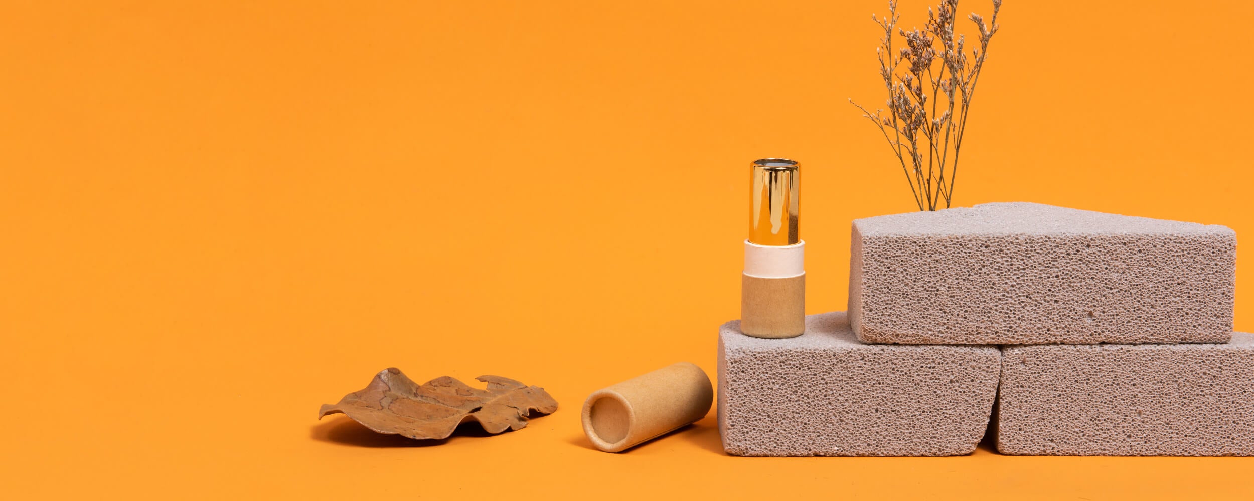 A kraft twist-up paper tube with a metal cap stands atop terrazzo blocks beside a second tube, dried botanicals, and wood pieces on an orange background.