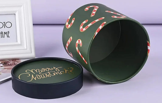 A green cylindrical container adorned with candy cane designs lies open on its side next to a lid that reads "Merry Christmas" in gold text, reflecting elegant holiday packaging, placed on a surface near a white picture frame.