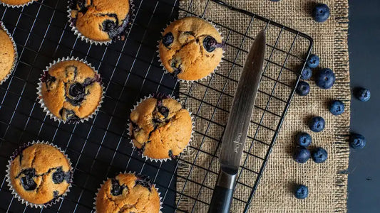 Blueberry muffins cooling on a black wire rack, with a knife and scattered fresh blueberries on a burlap cloth beside them, evoke the perfect morning scene.