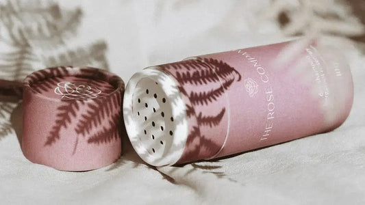 A cylindrical pink paper container with a perforated lid lies open on a soft fabric surface with fern shadows cast across it.