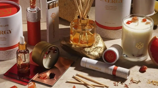 A variety of wellness and aromatherapy products, including a candle, diffuser, essential oils, and pretty cosmetic packaging, are displayed on a surface with pomegranate seeds scattered around.