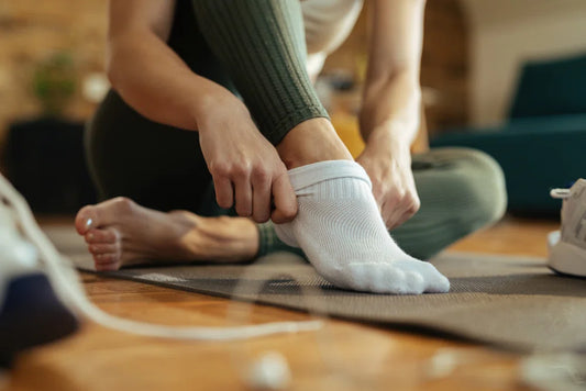 Close-up of a person putting on white socks while sitting on a yoga mat, with one foot already wearing a sock and the other barefoot, sneakers nearby.