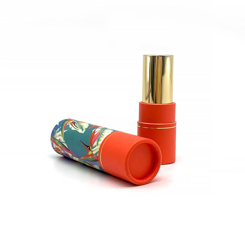 A vibrant paper tube with a tropical print lies next to an open lipstick with a glossy gold cap and a bold orange base, against a white backdrop.