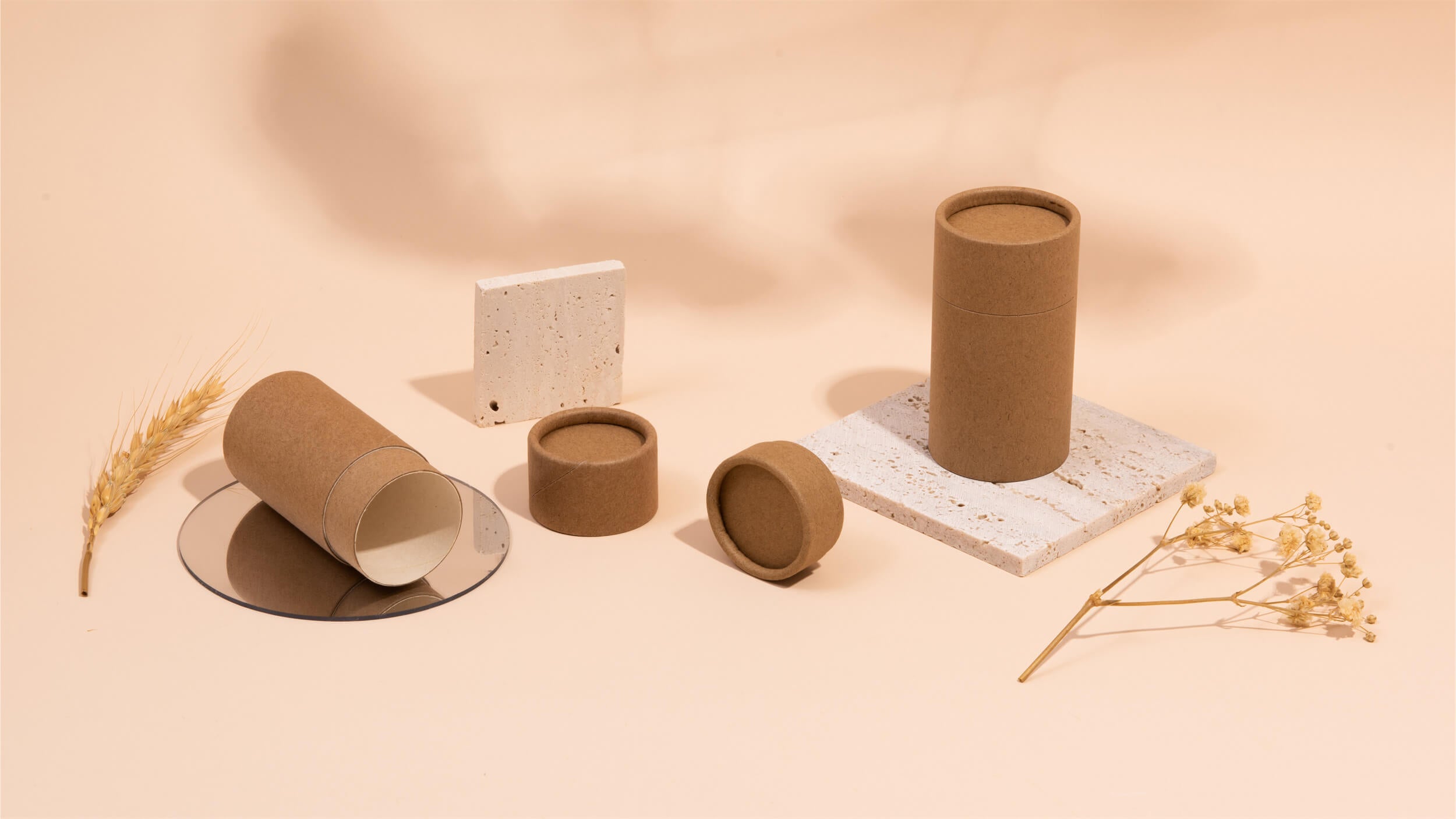 Varied sizes of cylindrical kraft paper tubes casually arranged on a peach background with white terrazzo blocks, a stalk of wheat, and dried flowers for decoration.