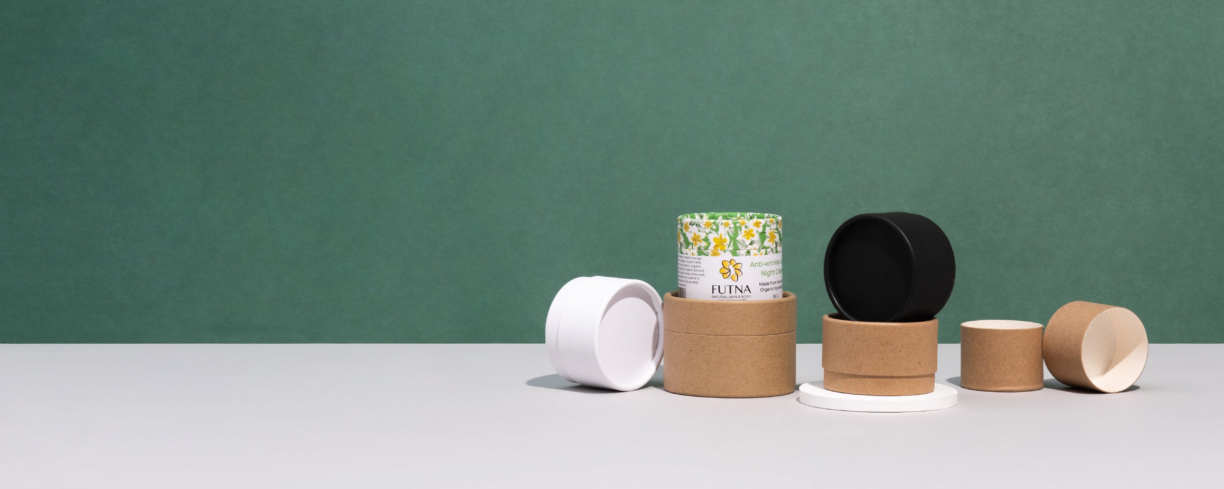 An array of cylindrical paper tubes in white and natural cardboard colors, with one featuring a floral design with customized logo, arranged on a two-tone surface against a green backdrop.