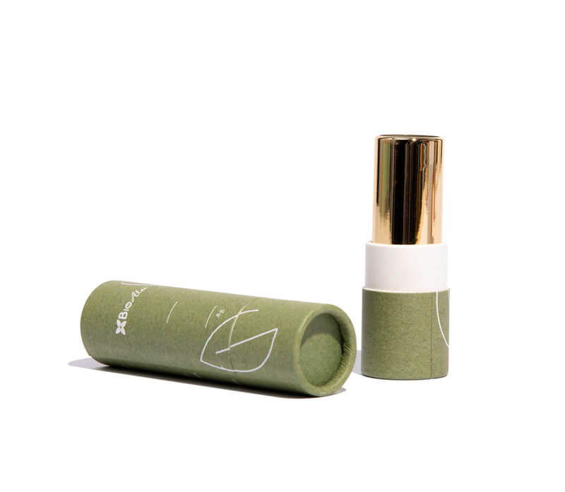 A green paper tube with white line drawings lies next to a cosmetic bottle with a golden cap and white label, indicating natural or organic beauty product packaging.