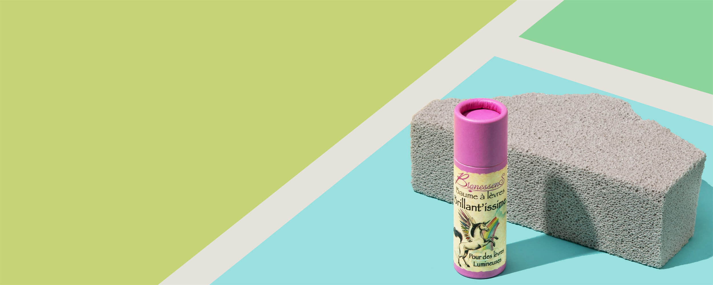 A colorful paper tube with a vibrant pink cap rests beside a textured gray stone on a split background of mint green and pastel blue.