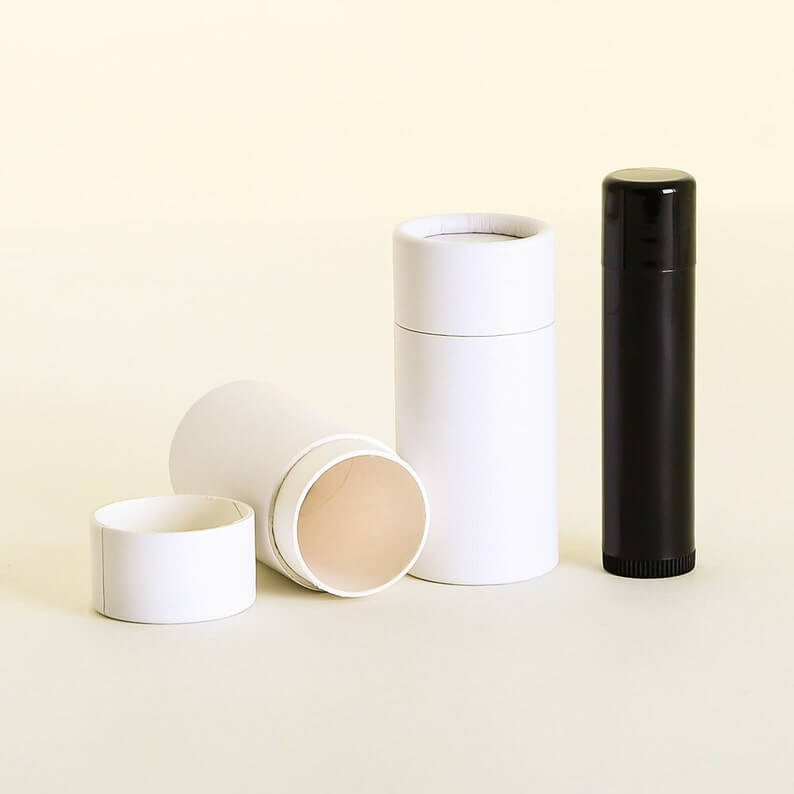 Three white cylindrical containers with one open lid, alongside a black tube, against a light beige background.